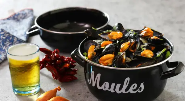 Un bicchiere di birra dorata accanto a cozze fumanti in una pentola nera, con peperoncini secchi e scalogni. Un perfetto abbinamento di birra e pesce.