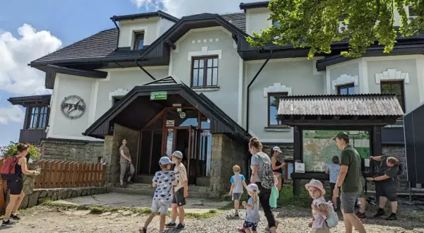 Rodzina przy schronisku na Magurce Wilkowickiej. Dzieci zwiedzają okolicę, a dorośli planują trasę na mapie.