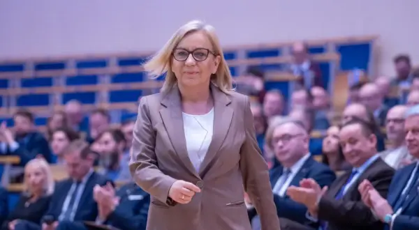 Kobieta w okularach i brązowej marynarce idzie na tle publiczności.