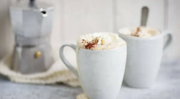 Två muggar med krämig mocca kaffe, toppad med vispgrädde och kakao. En mokabryggare står i bakgrunden, redo för ett nytt mocca kaffe recept.