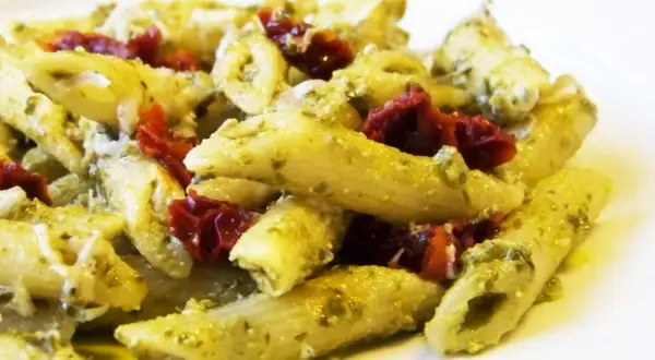 Makaron penne z zielonym pesto i suszonymi pomidorami na białym talerzu.
