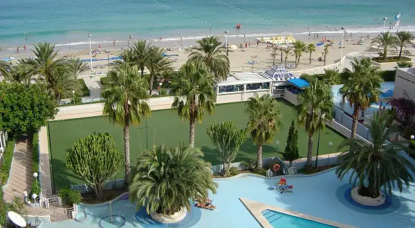 Vista aérea de un resort con piscina, cancha de césped, palmeras y una playa con el mar azul. Un lugar perfecto para sol y mar spa.