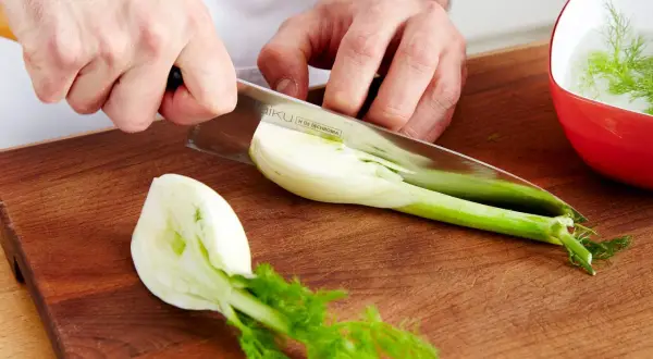 Hände schneiden Fenchel für leckere Fenchel Rezepte. Ein Teil liegt auf dem Holzbrett, ein anderer in einer roten Schüssel mit Wasser.