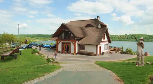 Dom z piękną, naturalną strzechą na dachu, położony nad jeziorem. Wokół zielony trawnik, drewniany wóz i rzeźby.