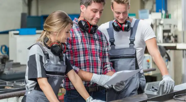 Drei SHK-Techniker besprechen Pläne. Sie tragen Arbeitskleidung und Kopfhörer. Das Gehalt eines SHK-Technikers ist ein wichtiges Thema.