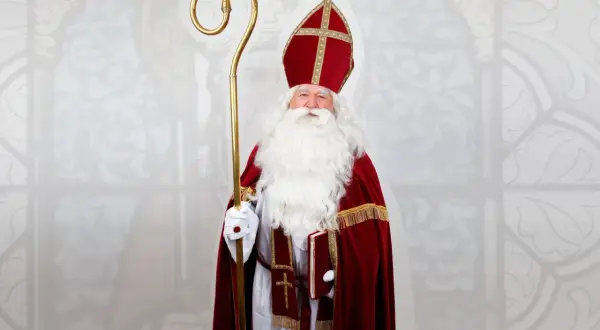 Mężczyzna w stroju św. Mikołaja biskupa, z białą brodą, mitrą i pastorałem, trzyma książkę.