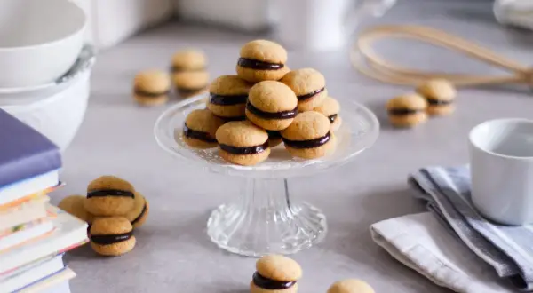 Baci di dama, deliziosi biscotti con crema al cioccolato, disposti su un'alzata di vetro, circondati da libri e tazze.