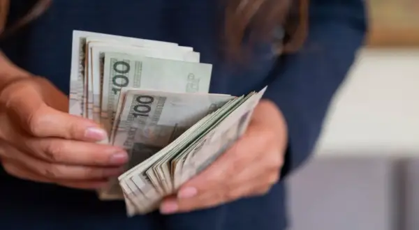 Dłonie trzymające plik banknotów 100 zł. Może to symbolizować wynagrodzenie lub zarobki w budżetówce, gdzie grupy zaszeregowania wpływają na pensję.