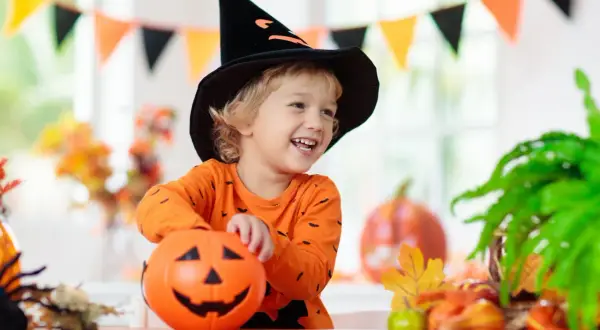Uśmiechnięty chłopiec w czarnej, spiczastej czapce czarownicy i pomarańczowej bluzce z nietoperzami trzyma dynię. To idealny strój owoc dla dziecka na Halloween!