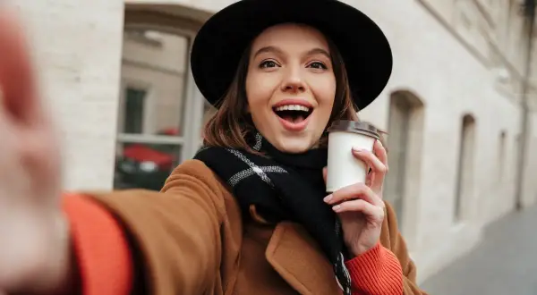 Uśmiechnięta dziewczyna w kapeluszu i płaszczu robi selfie z kawą. Chcesz wiedzieć, jak ustawić makijaż w aparacie iPhone?