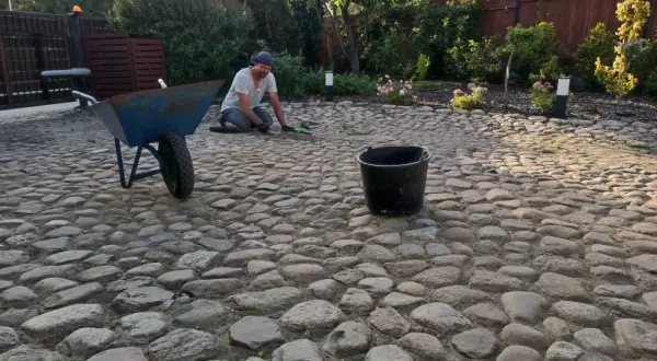 Mężczyzna pracuje przy podjeździe z kamienia polnego, obok stoi taczka i wiadro.