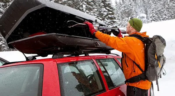Mann lädt Skier in eine offene Dachbox auf einem roten Auto im Schnee.