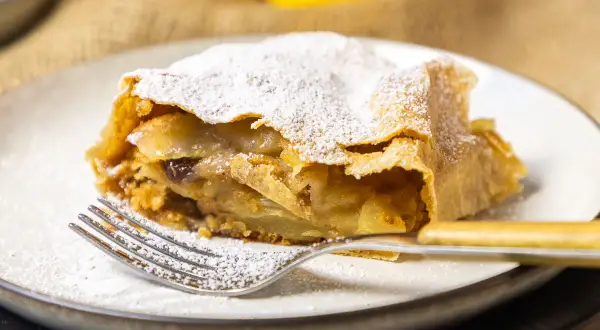 Ein Stück Apfelstrudel, bestäubt mit Puderzucker, wartet auf dem Teller. Dieses Apfelstrudel Rezept ist ein Genuss!