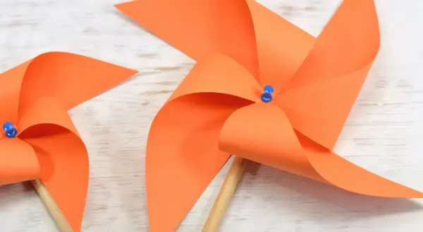 Zwei selbstgebastelte Windräder aus orangefarbenem Papier auf Holzuntergrund. Perfekt zum Windrad basteln mit Kindern.