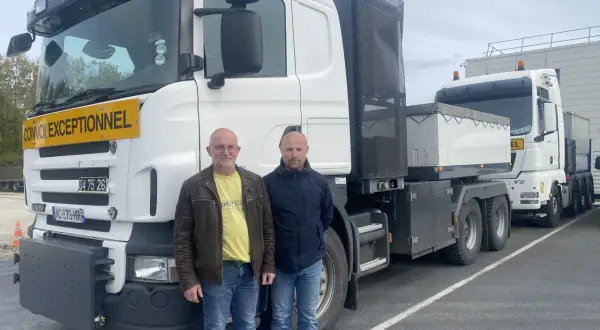 Deux chauffeurs poids lourd national posent devant leurs camions. L'un porte un gilet jaune, l'autre une veste bleue.