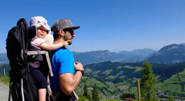 Vater trägt sein Baby in einer Kraxe auf einem Berg. Die Aussicht ist atemberaubend.