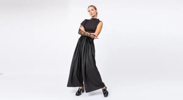 Kobieta w czarnym, eleganckim stroju z szerokimi spodniami i siateczkowymi rękawami, pozuje na białym tle.