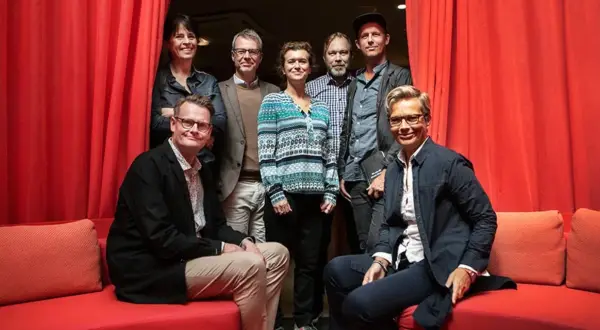 Gruppbild med åtta personer framför röda draperier. Izabella Jensen står i mitten, klädd i en mönstrad tröja.