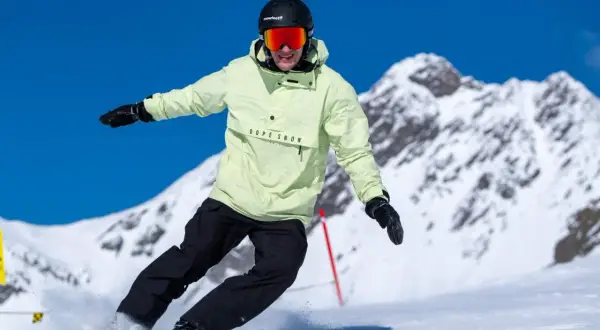 Snowboarder w jasnej kurtce i czarnych spodniach, z kaskiem i goglami, śmiga po stoku. Czy buty narciarskie pasują do snowboardu? Nie, to różne sprzęty.
