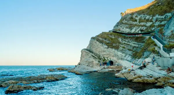 Scogliere rocciose e mare blu intenso al Conero, con un tunnel scavato nella roccia e persone che si godono la giornata.
