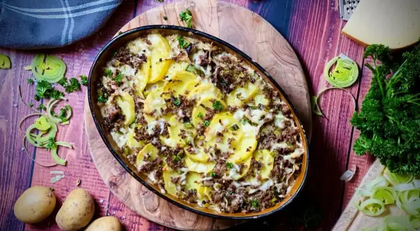 Herzhafter kartoffelauflauf mit hackfleisch, überbacken mit Käse und garniert mit Petersilie. Dazu frische Kartoffeln, Lauch und ein Stück Käse.