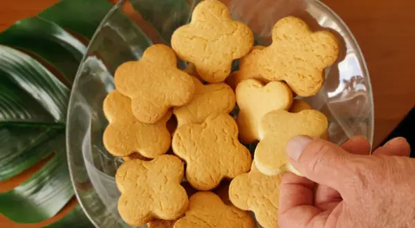 Una mano prende uno dei tanti biscotti fatti in casa a forma di quadrifoglio, disposti in una ciotola di vetro con foglie verdi sullo sfondo.