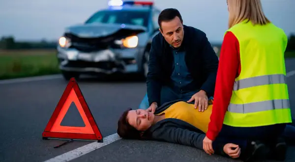 Un hombre y una mujer asisten a una persona en el suelo tras un accidente. Un triángulo de señalización y un coche dañado en el fondo ilustran la importancia de la seguridad vial.