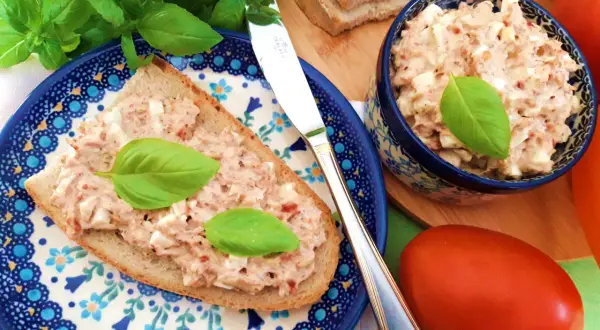 Pyszna pasta jajeczna z tuńczykiem na kromce chleba, ozdobiona bazylią. Obok miseczka z pastą i świeże pomidory.