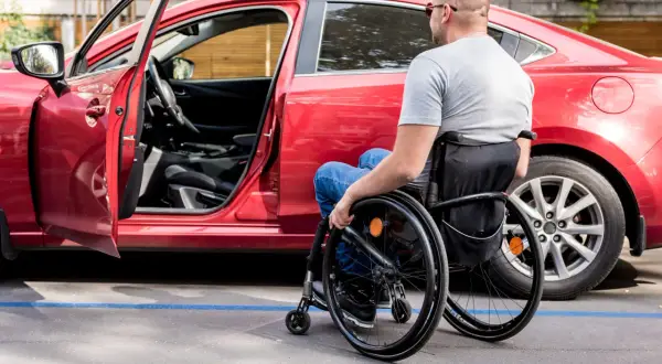 Uomo in carrozzina accanto a un'auto rossa, con portiera aperta. Un esempio di accessibilità, come previsto dall'art. 30 comma 7 legge 388 del 2000.