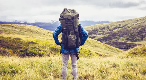 Wanderer mit großem Rucksack steht auf einem Hügel, bereit für seine 1 Jahr Weltreise. Die Landschaft ist grün und hügelig.