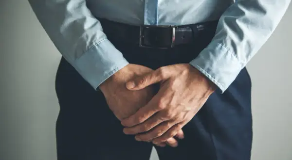 Un homme en chemise bleue et pantalon sombre, mains croisées devant lui, semble avoir un testicule qui gratte.