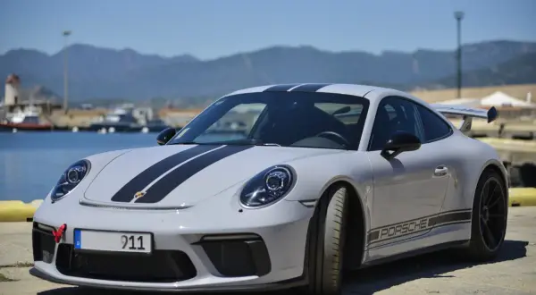 Szary Porsche Carrera 911 z czarnymi pasami na masce i dachu, zaparkowany przy nabrzeżu z widokiem na góry.