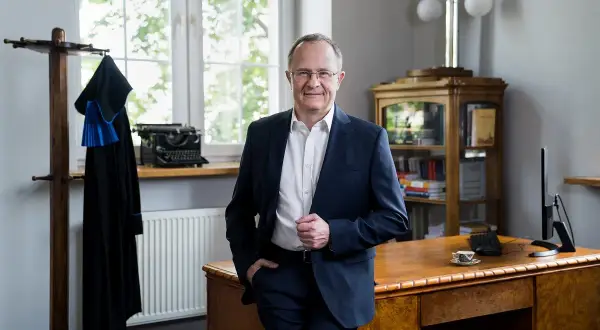 Mężczyzna w garniturze stoi w gabinecie, obok wieszaka z togą prawniczą i zabytkowej maszyny do pisania.