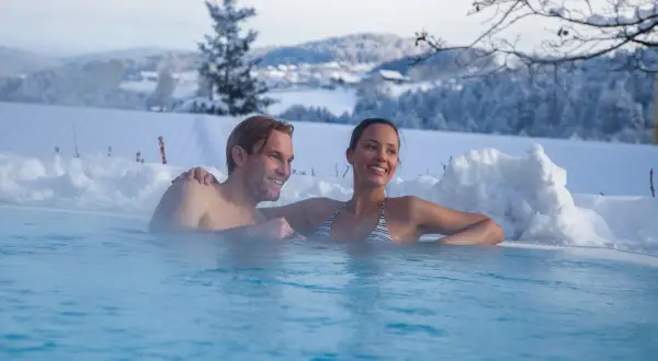 Ein Paar entspannt in einem beheizten Pool im Schnee mit verschneiter Landschaft im Hintergrund.