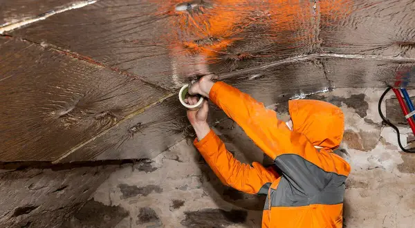 Travaux d'isolation dans une cave en pierre. Un ouvrier en combinaison orange fixe un isolant réfléchissant au plafond avec du ruban adhésif.