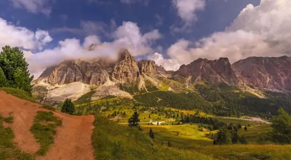 Camino de tierra rojiza que se adentra en un valle alpino con imponentes montañas cubiertas de nubes. La ruta Cinque Torri se revela entre prados verdes y bosques frondosos.