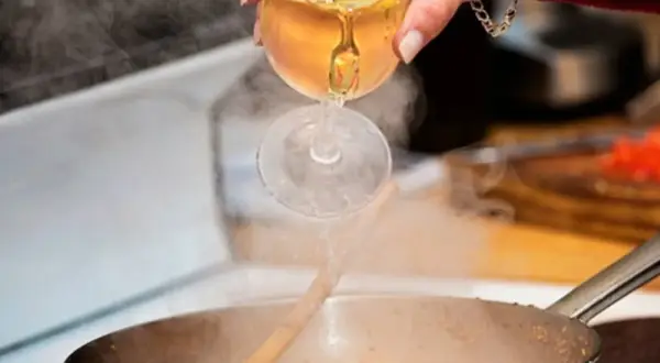 Eine Hand gießt Weißwein aus einem Glas in eine dampfende Pfanne. Das Verkaufen von Wein beim Kochen verleiht dem Gericht Tiefe.