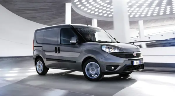 Szary Fiat Doblo Cargo w ruchu, z rozmytymi kołami, na tle nowoczesnej architektury z okrągłymi światłami.