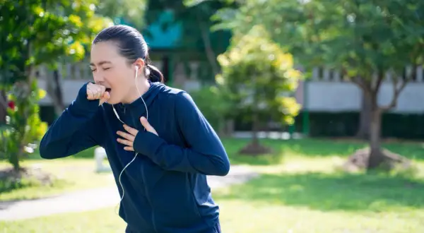 Donna con cuffie che corre in un parco, colpita da un attacco di tosse, si porta una mano alla gola.
