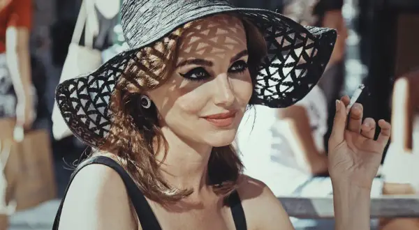Kobieta w kapeluszu z lat 60. i wyrazistym makijażu oczu pali papierosa.
