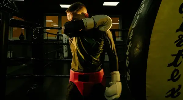 A boxer wipes sweat from his face in a dimly lit boxing ring. He's wearing gloves and a red cup. This image makes you wonder how long do boxers rest after a fight.