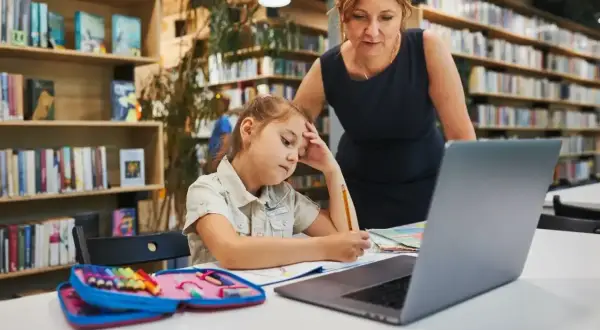 Dziewczynka z ołówkiem w dłoni, zamyślona nad zadaniem, przygotowuje podanie do szkoły średniej. Nauczycielka wspiera ją w bibliotece.
