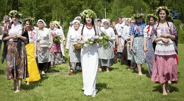 Grupa kobiet w tradycyjnych strojach i wiankach idzie przez łąkę. Centralna postać w białej sukni niesie bukiet kwiatów. Obraz nawiązuje do tradycji Zielonych Świątek.