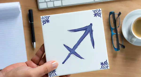 Dłoń trzyma kwadratową płytkę z symbolem Strzelca, znakiem zodiaku dla osób urodzonych 28 listopada.