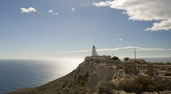El faro de Mesa Roldán se alza sobre un acantilado rocoso, vigilando el mar Mediterráneo bajo un cielo azul con nubes dispersas.