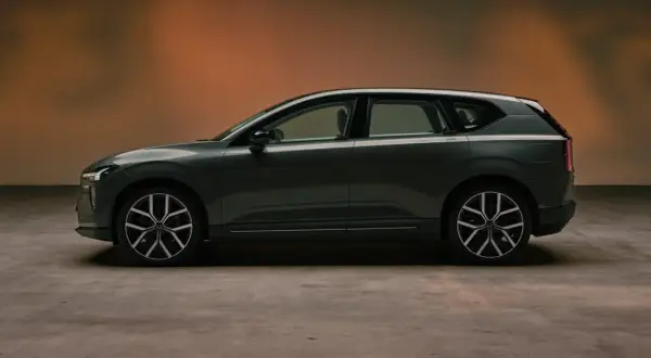 Volvo EX60 gris oscuro, perfil lateral, con fondo degradado naranja y suelo de hormigón.