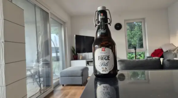 Eine Flasche Moritz Fiege Pils steht auf einem Tisch. Im Hintergrund ein Wohnzimmer mit Sofa und Fernseher.