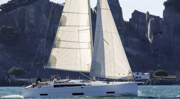 Un elegante catamarano solca acque azzurre, con vele bianche gonfie di vento, mentre sullo sfondo si stagliano scogliere rocciose.