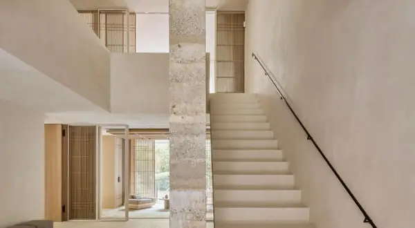 Escaleras de cemento blancas y minimalistas ascienden junto a un pilar de piedra texturizada, conectando niveles en un interior luminoso.