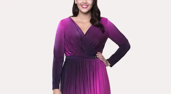 Uśmiechnięta modelka w butik na plus sukienki. Długa, fioletowo-różowa sukienka z dekoltem kopertowym i długimi rękawami.
